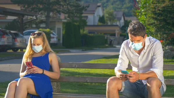 CERRAR: Dos extraños usan mascarillas mientras están sentados en el banco del parque y enviando mensajes de texto. —  Fotos de Stock