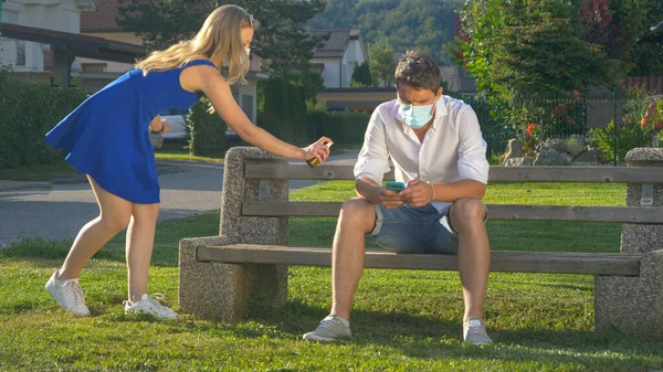 CLOSE UP: Mädchen mit Gesichtsmaske sprüht Mann, der auf Bank sitzt und SMS schreibt. — Stockfoto