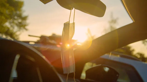 CLOSE UP, DOF: Surgical mask hangs off a rear-view mirror inside a sunlit car. — Stock Photo, Image