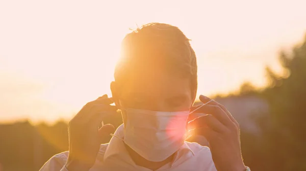 CLOSE UP: Smiling Caucasian man puts on a disposable facemask at golden sunset. — Stock Photo, Image