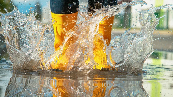 CLOSE UP Unrecognizable girl in yellow rubber boots jumps into the glassy puddle