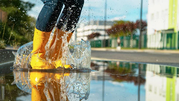 LOW ANGLE: Girl in bright yellow rubber boots jumps into the glassy puddle. — Stock Photo, Image