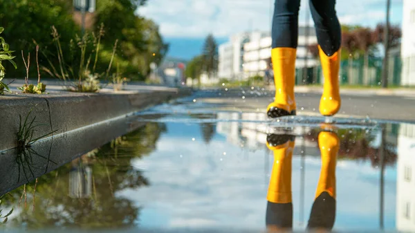 LOW ANGLE: Розмита жінка в жовтих дощових черевиках біжить у велику скляну калюжу . — стокове фото