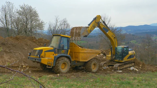 Big excavator unloads a bucket full of soil in the back of a yellow truck. — Stock Photo, Image