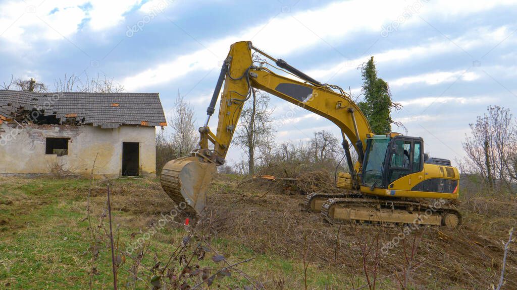 Yellow excavator clears branches off a meadow near the ruins of an old house