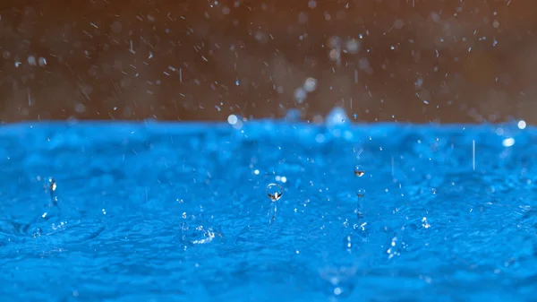 MACRO: Drops of rainwater splatter upon contact with the light blue pool water.