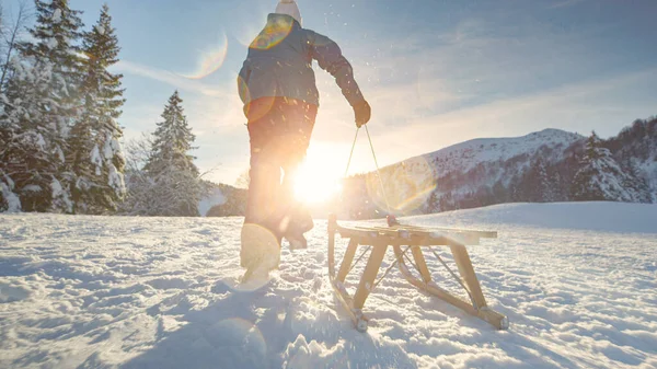 LOW ANGLE: Unrecognizable woman runs up a snowy hill with her wooden sleigh. — Stock Photo, Image