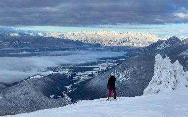 Genç snowboardcu bir dağın tepesinde duruyor ve nefes kesici manzarayı gözlemliyor.
