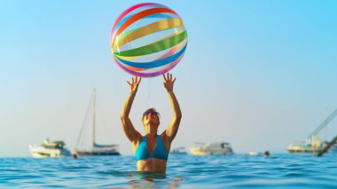 Bikinili genç bir kadın güneşli bir akşamda kocaman şişme bir topla oynuyor.