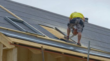 İnşaatçı bitmemiş bir çatının tepesinde duruyor ve tahtadan bir açıklık kesiyor..