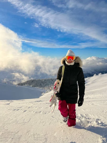 ZAMKNIJ Uśmiechnięta młoda kobieta wspina się na niezadbane wzgórze podczas jazdy na snowboardzie — Zdjęcie stockowe