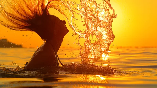 CLOSE UP: Woman splashes glassy ocean water with her hair at golden sunset.