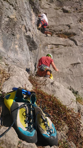 VERTICAL: Os sapatos de alpinista estão no chão enquanto outros escalam a parede rochosa. — Fotografia de Stock