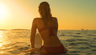Tanımlanamayan sörfçü kız sörf tahtasına oturur ve günbatımını izler.