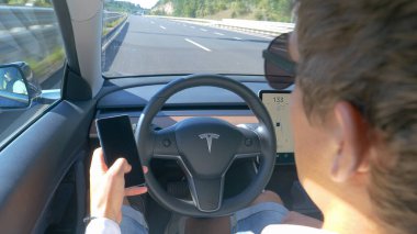 Tesla 'nın arabasında oturan banliyö çalışanı otoyolda giderken telefonu kontrol ediyor.