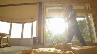 LoW ANGLE Builder omuzunda ahşap bir panel ile güneşli bir odada yürür