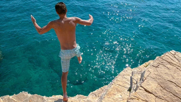 Carefree man dives off a towering rocky cliff and into the deep blue sea. — Stockfoto