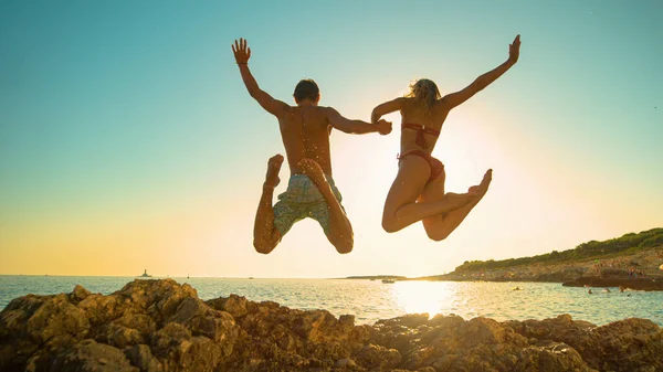 ÁNGULO BAJO: Alegre pareja joven toma de la mano mientras se sumerge en el océano al atardecer — Foto de Stock