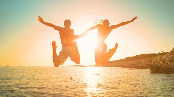 LENS FLARE: Pareja alegre en vacaciones de verano salta al océano al amanecer. — Foto de Stock