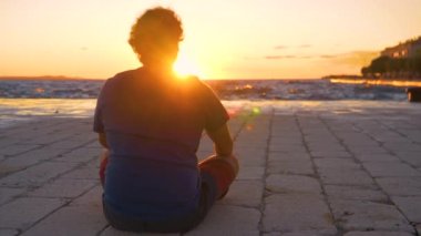 Genç adam taş zeminde oturur ve gün batımında denizi izler..