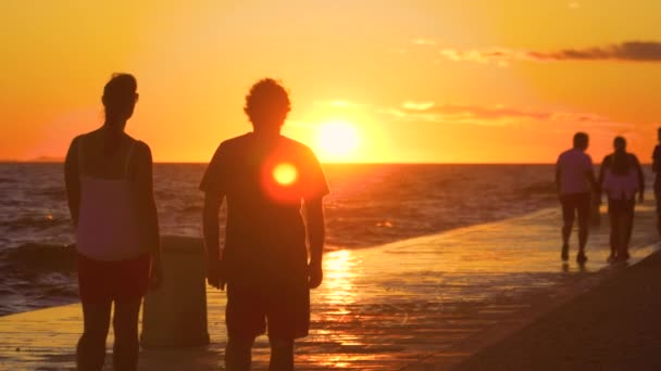 SILHOUETTE:若い女性と男性が夕日の美しい海岸沿いの遊歩道を歩く. — ストック動画