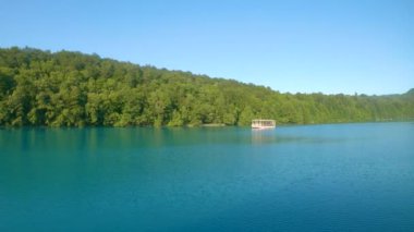 Turist teknesi Plitvice 'deki zümrüt göl boyunca dolaşır ve yemyeşil ormanlarla çevrilidir..