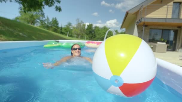 CLOSE UP: Smiling woman swims towards an inflatable ball floating in her pool. — стоковое видео