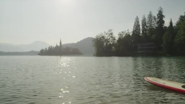 MOCIÓN LENTA: Chicas surfistas jóvenes SUPing hacia la isla del lago Bled al amanecer — Vídeo de stock