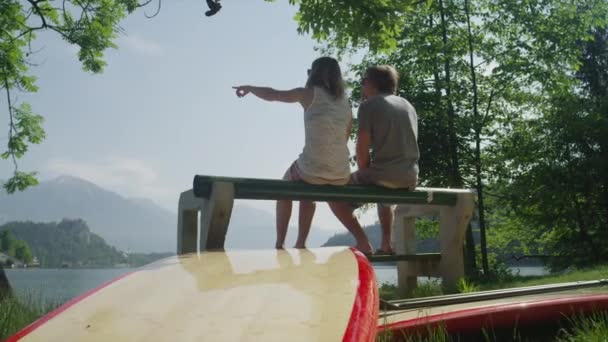 MOTION SLOW : Jeune couple assis sur un banc au bord du lac avec des planches SUP parler — Video