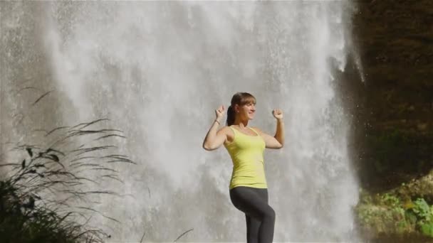 Веселая молодая женщина поднимает руки перед водопадом — стоковое видео