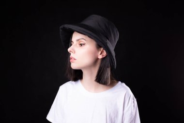 Beyaz tişörtlü, siyah panama şapkalı, siyah arka planda portresi olan hoş bir kadın.