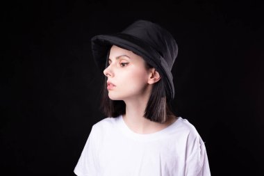 Beyaz tişörtlü, siyah panama şapkalı, siyah arka planda portresi olan hoş bir kadın.