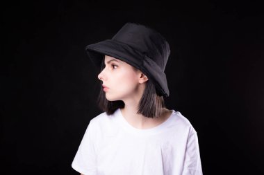 Beyaz tişörtlü, siyah panama şapkalı, siyah arka planda portresi olan hoş bir kadın.
