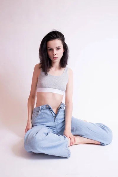 woman in blue jeans and sports top sits, white studio background