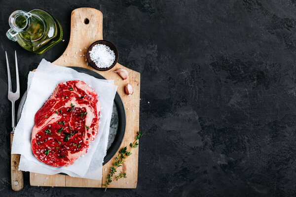 Raw beef steak on cutting board with salt and seasonings. Fresh steak meat prepared for cooking on a dark stone background, top view, copy space.