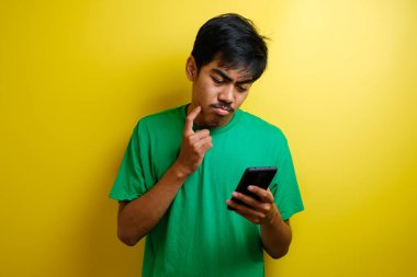 Genç Asyalı adam gülümsüyor, düşünüyor ve yukarı bakarken akıllı telefonuyla mesajlaşıyor, sarı arka planda yarı vücut portresi.