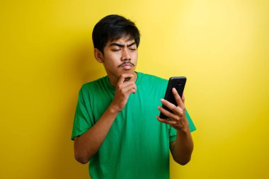 Genç Asyalı adam gülümsüyor, düşünüyor ve yukarı bakarken akıllı telefonuyla mesajlaşıyor, sarı arka planda yarı vücut portresi.