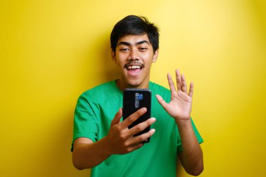 Asyalı bir adam selfie çekiyor ya da evde video görüşmesi yapıyor kameraya gülümsüyor ve el sallıyor. Sarı arka planda neşeli bir ifade