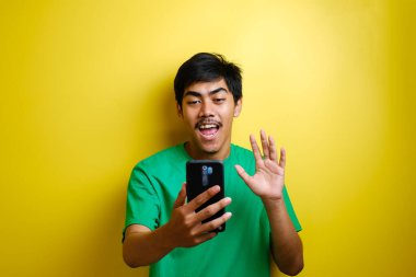 Asyalı bir adam selfie çekiyor ya da evde video görüşmesi yapıyor kameraya gülümsüyor ve el sallıyor. Sarı arka planda neşeli bir ifade