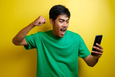 Çekici genç Asyalı adamın telefonunda iyi haberler var. Sarı arka plana karşı gülen mutlu bir gülücük.