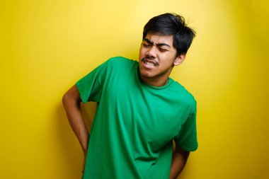 Yeşil tişört giyen, sırt ağrısı çeken, sırtını ovuşturan, yorgun bir Asyalı genç adam. Sarı arkaplan