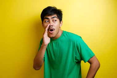 Yeşil tişörtlü genç Asyalı bir adamın portresi. Gizlice konuşuyor. Sarı arka plandaki birine sırrını anlatıyor.