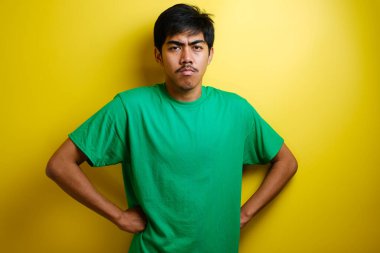 Asyalı genç adam alaycı mutsuz surat ifadesi sergiliyor, kameraya bakıyor, elleri kalçasında, sarı arka planda izole edilmiş.