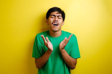 Yeşil tişörtlü genç Asyalı adam kolunu kaldırırken gözlerini kapat göğsü üzgün görünüyor, sarı arka plana karşı başarısız bir hareket sergiliyor.
