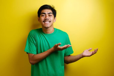Gülümseyen yakışıklı Asyalı adam sarı arka planda izole edilmiş boş bir eli işaret ediyor.