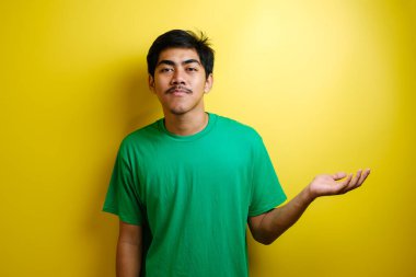 Gülümseyen yakışıklı Asyalı adam sarı arka planda izole edilmiş boş bir eli işaret ediyor.