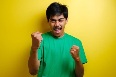 Heyecanlı genç Asyalı adam, gülümseyen mutlu yüzüyle yumruklarını kaldırıyor, evet jest, sarı arka planda başarıyı kutluyor.