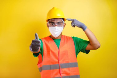 Asyalı inşaat işçisi koruyucu maskesini ve kurnaz miğferini göstererek sarı arka plana karşı sağlığa ve güvenliğe öncelik veriyor.