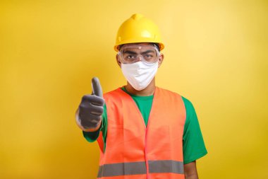 Asyalı inşaat işçisi koruyucu maskesini ve kurnaz miğferini göstererek sarı arka plana karşı sağlığa ve güvenliğe öncelik veriyor.