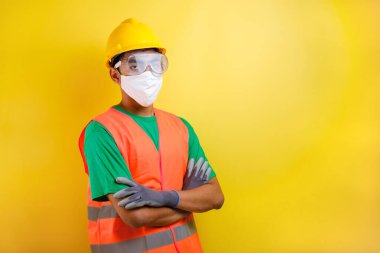 Maskeli ve güvenlik gözlükleri takan Asyalı müteahhit, sarı arka plana karşı güven jesti yaparak kolunu çarpmış.